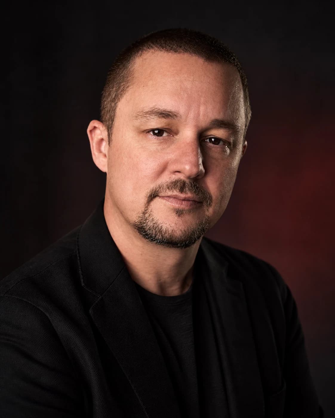 Portrait of Tony Simons, host of Less Lip, More Ship, in a dark jacket against a dark red background